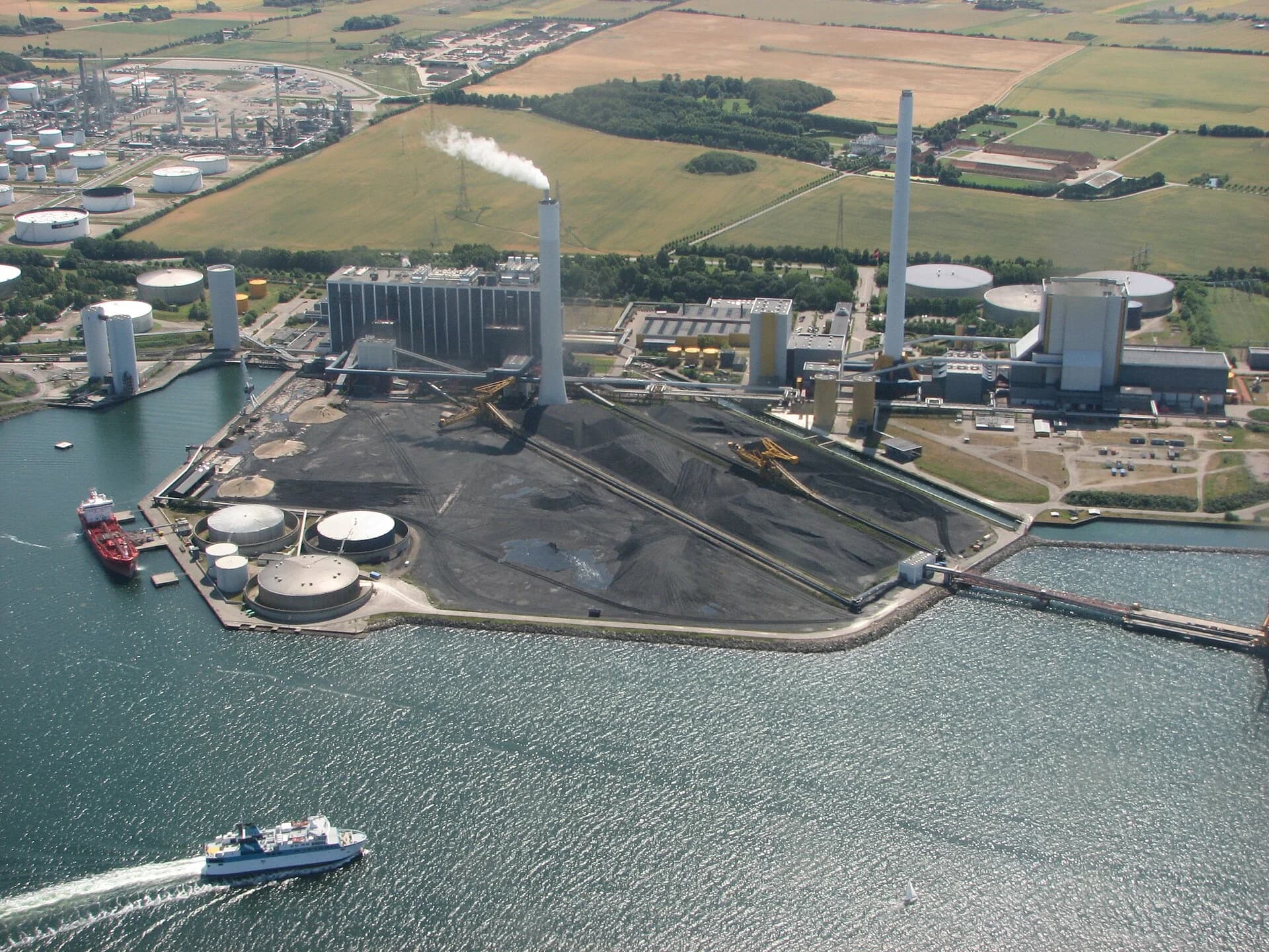 Aerial photograph of Asnæs power station at Kalundborg, Denmark, on the coast, with its tall chimneys, a large fuel stockpile, storage domes and a jetty, and the oil refinery’s storage tanks in the background.