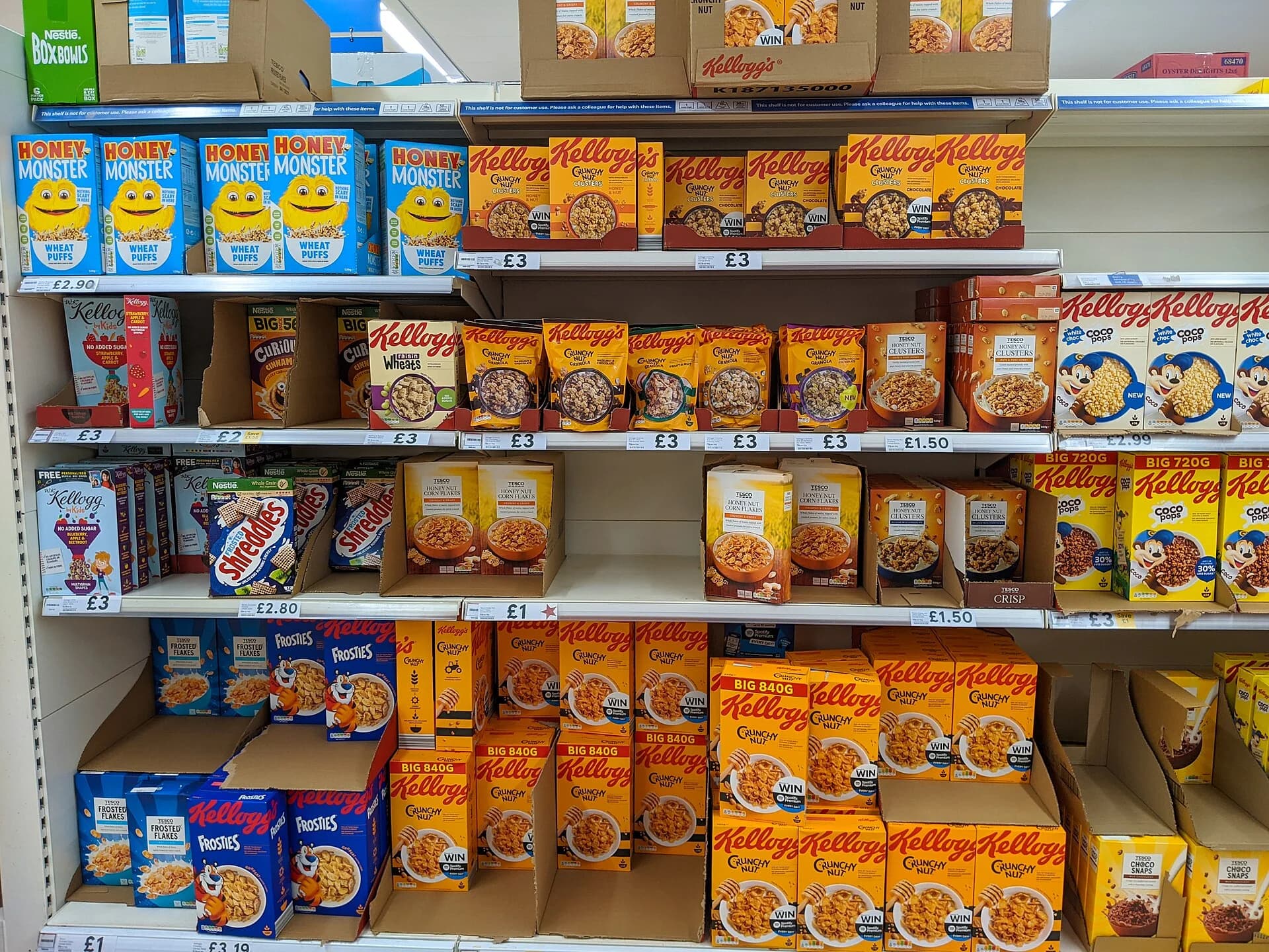 Supermarket breakfast cereal shelves filled mostly with Kellogg's brands (Crunchy Nut, Coco Pops, Frosties) alongside a few Nestlé, Honey Monster and Tesco own-label boxes, in many flavours and pack sizes.