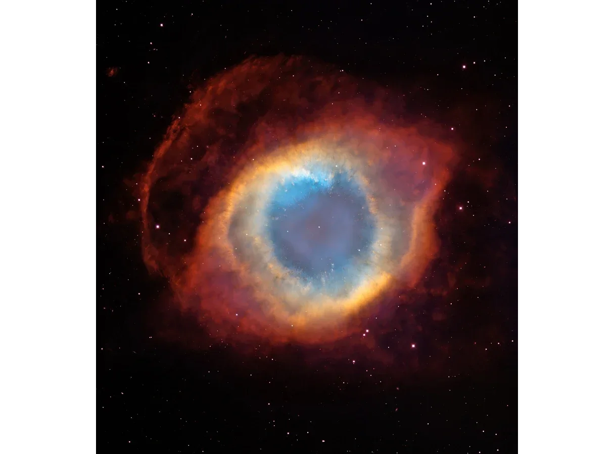 The Helix Nebula photographed by the Hubble Space Telescope and a ground-based telescope: a glowing ring of gas, blue-green inside and red at the edges, around a faint central star, against a black sky with background stars.