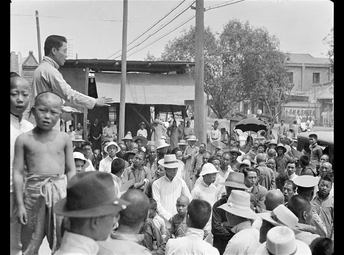 Black-and-white photograph of a street in Beijing. A student stands on a raised spot addressing a crowd of men in long gowns and caps; others stand and listen in the street.