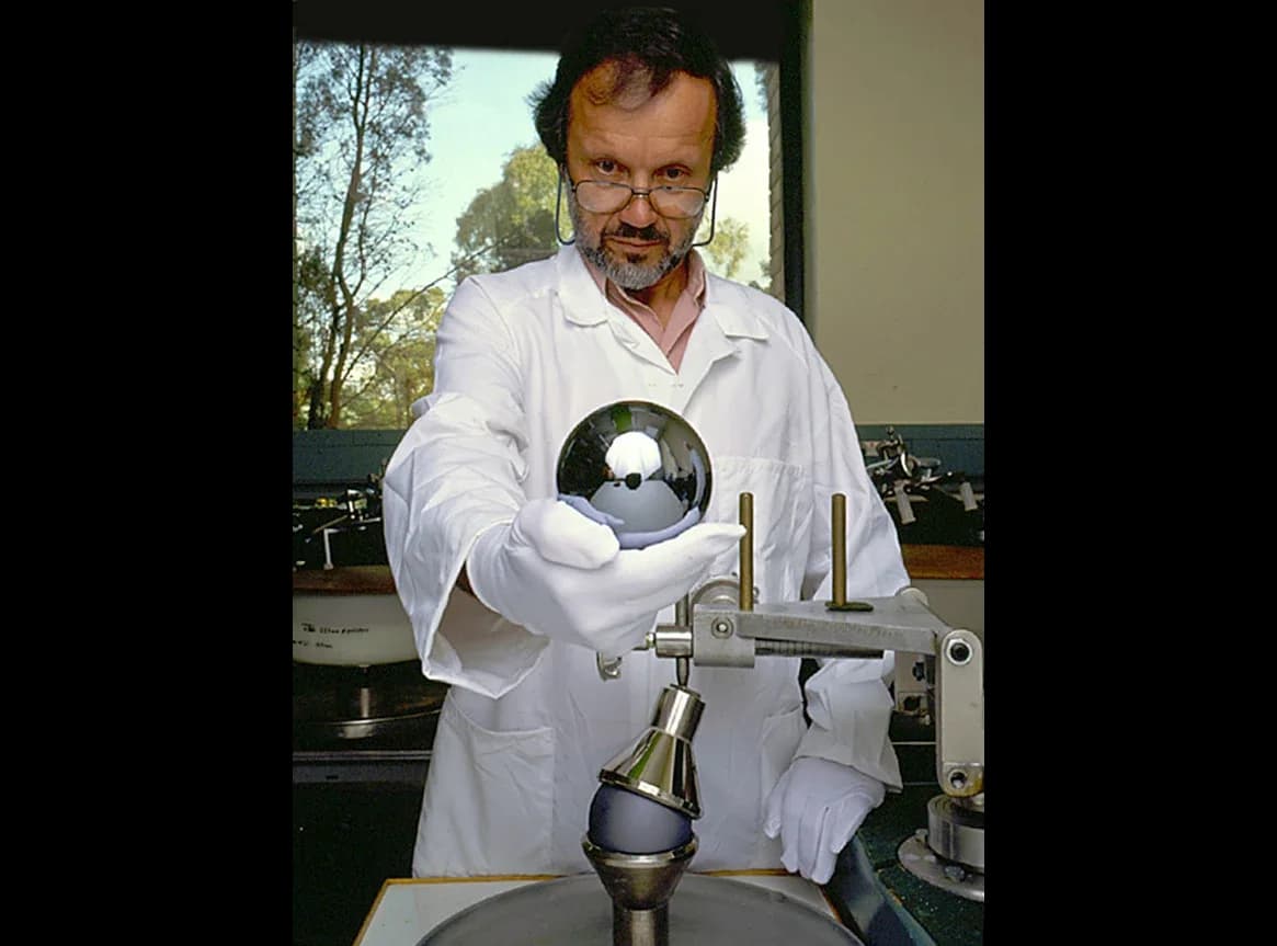 A highly polished, mirror-like sphere of single-crystal silicon about the size of a grapefruit, held up in two hands by an optician.