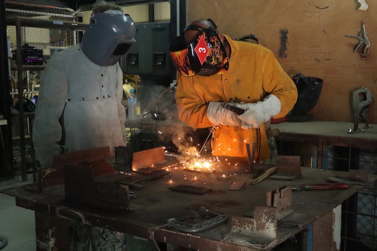 Two trainees in protective jackets and welding masks at a workbench in a training workshop; one welds a steel plate, sending up sparks, while the other watches.