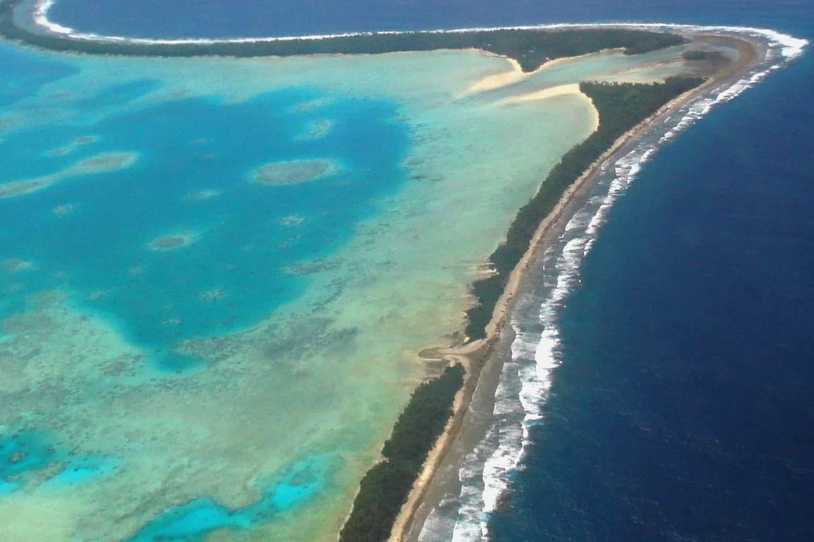 Aerial photograph of part of Funafuti atoll, Tuvalu: a narrow strip of land a few hundred metres wide, covered in trees and buildings, between the shallow turquoise lagoon on the left and the deep Pacific Ocean with breaking waves on the right.