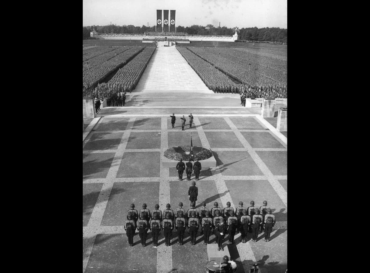 Black-and-white photograph taken from above: three men stand saluting before a wreath on a paved terrace, with a squad of helmeted men in the foreground and, beyond them, a wide stone path between huge blocks of uniformed men leading to a stand hung with three swastika banners.