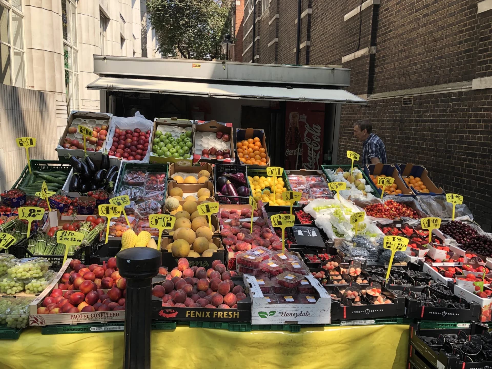 A London street fruit and vegetable stall with crates of apples, oranges, melons, peaches, berries and asparagus, each crate marked with a yellow price card such as £1 each, 3 for £1 or £4.00.