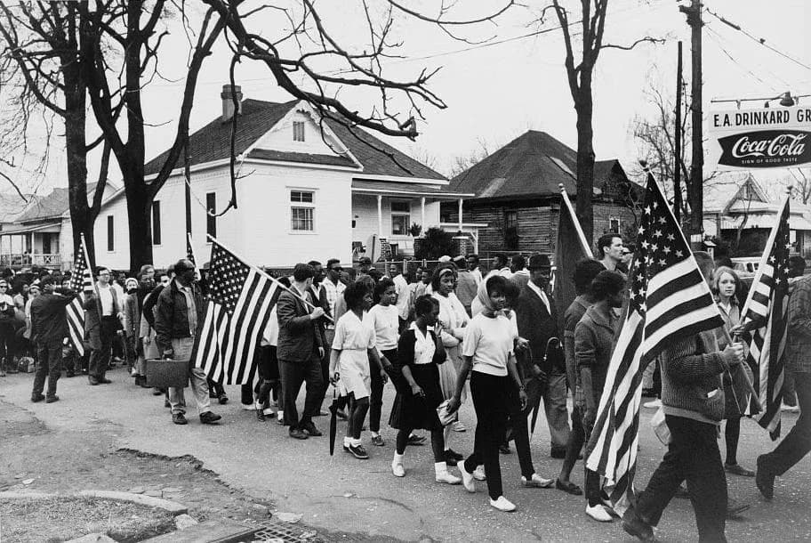 Black-and-white photograph of a long column of marchers walking along a road, several carrying American flags.