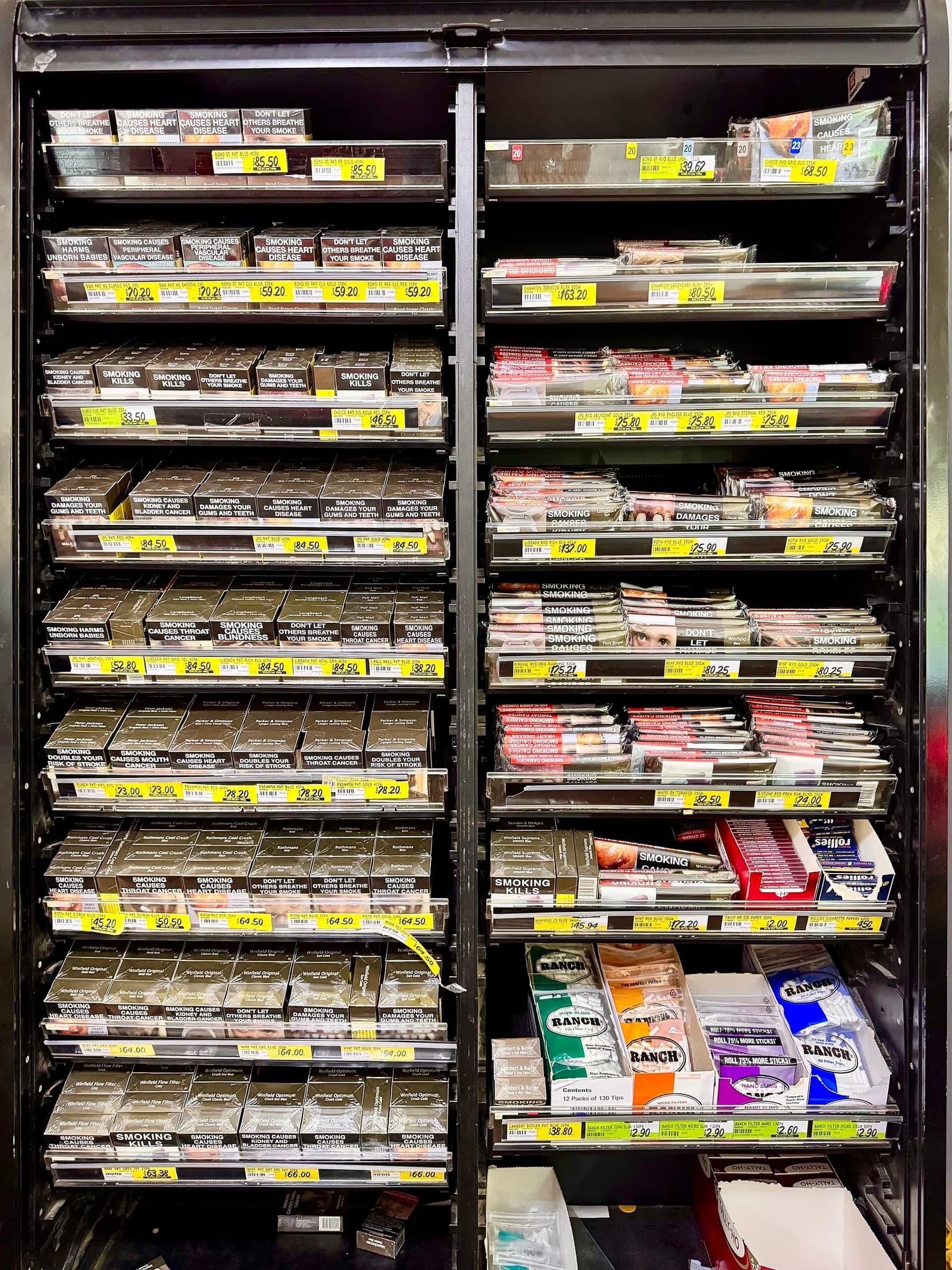 A shop's tobacco cabinet in Australia filled with plain olive-brown cigarette packs carrying large health warnings such as 'Smoking causes heart disease', with price labels on each shelf.