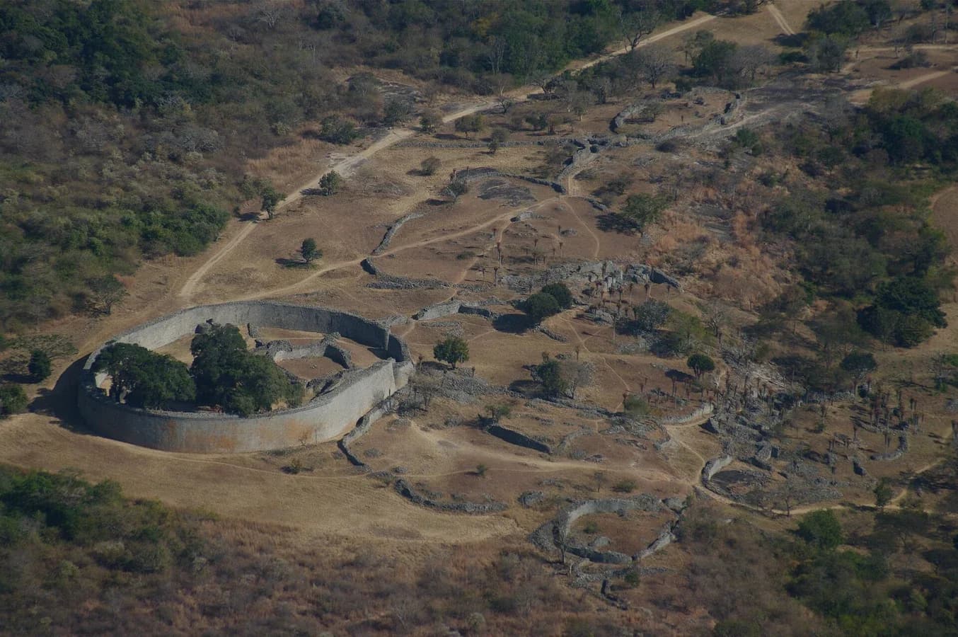 Photograph of the ruins of Great Zimbabwe taken from the hill above: a large oval enclosure of high dry-stone walls with trees growing inside, and lines of lower walled ruins stretching across the valley beyond it, surrounded by dry bush.