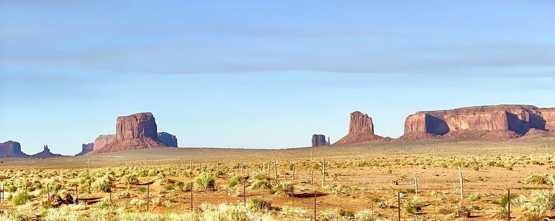 Monument Valley, Arizona: isolated red sandstone buttes with vertical cliffs and flat tops rise above a flat desert plain with scrub, and a broad flat-topped mesa stands on the right; aprons of debris slope from the foot of each cliff.