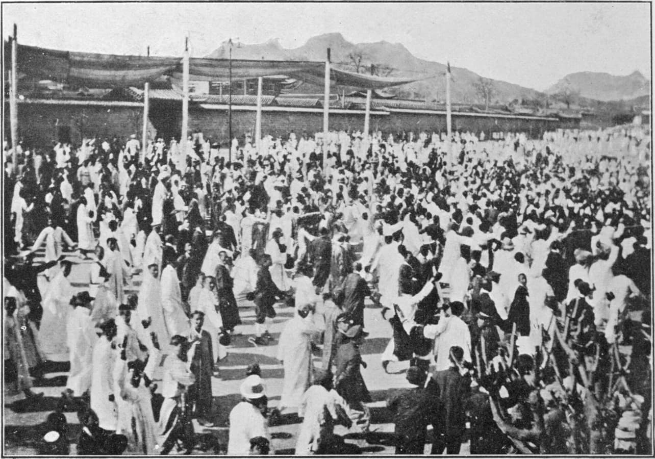 Black-and-white photograph of a large crowd of Korean people, most in white clothing and some raising their arms, gathered in an open space before a long wall in Seoul, with mountains behind.