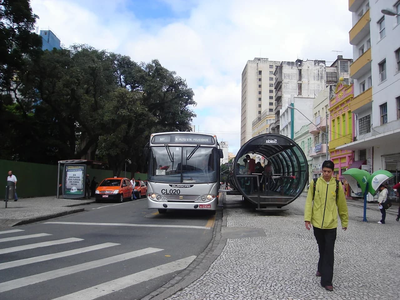 A Curitiba bus rapid transit stop: a glass tube station raised to bus-floor level beside a dedicated bus lane, with a bus approaching along a city-centre street.