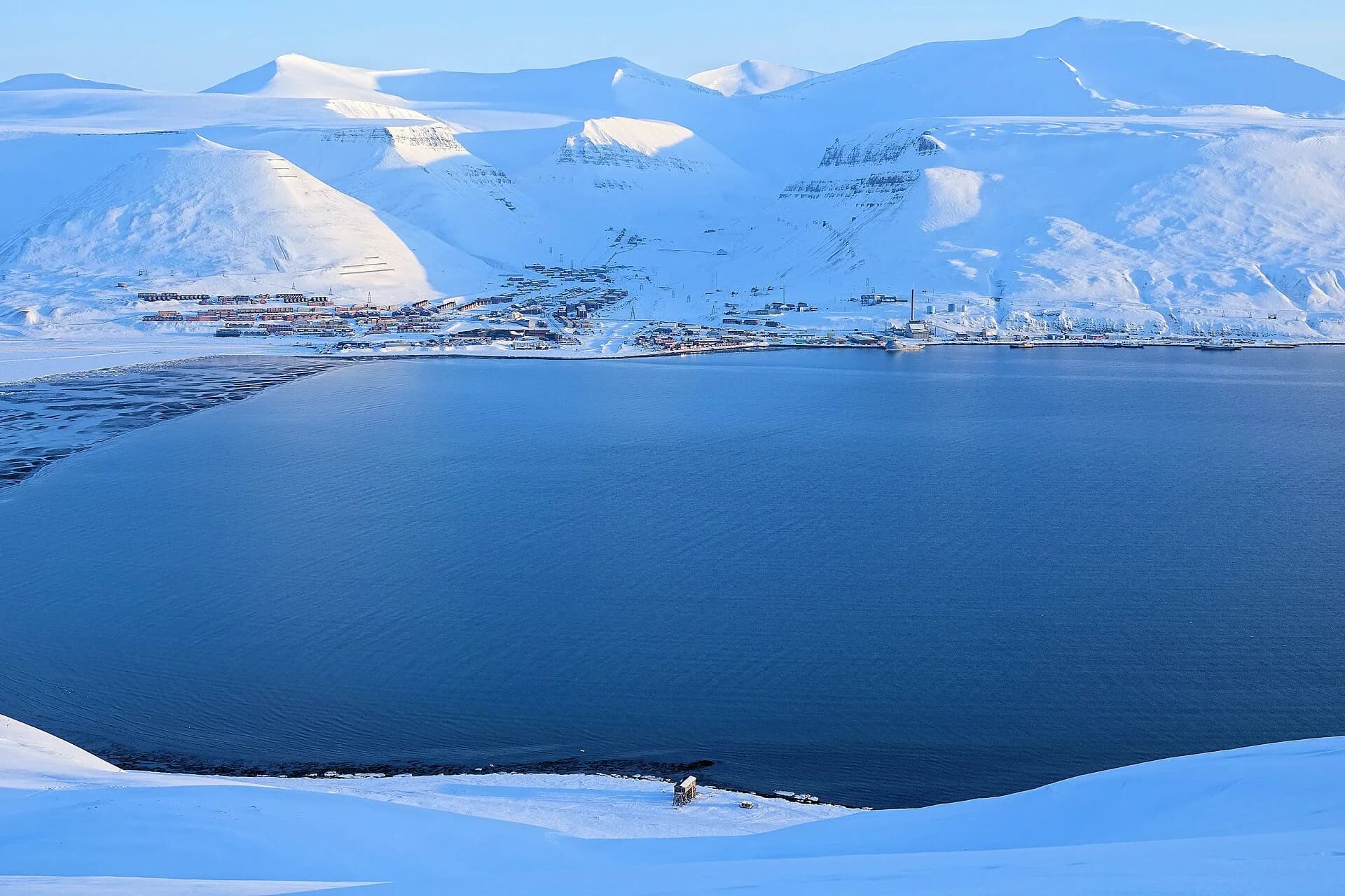 Longyearbyen seen across Adventfjorden in late winter: the town lies on a narrow strip of low ground at the mouth of a valley, with steep snow-covered mountain slopes rising directly behind the buildings.
