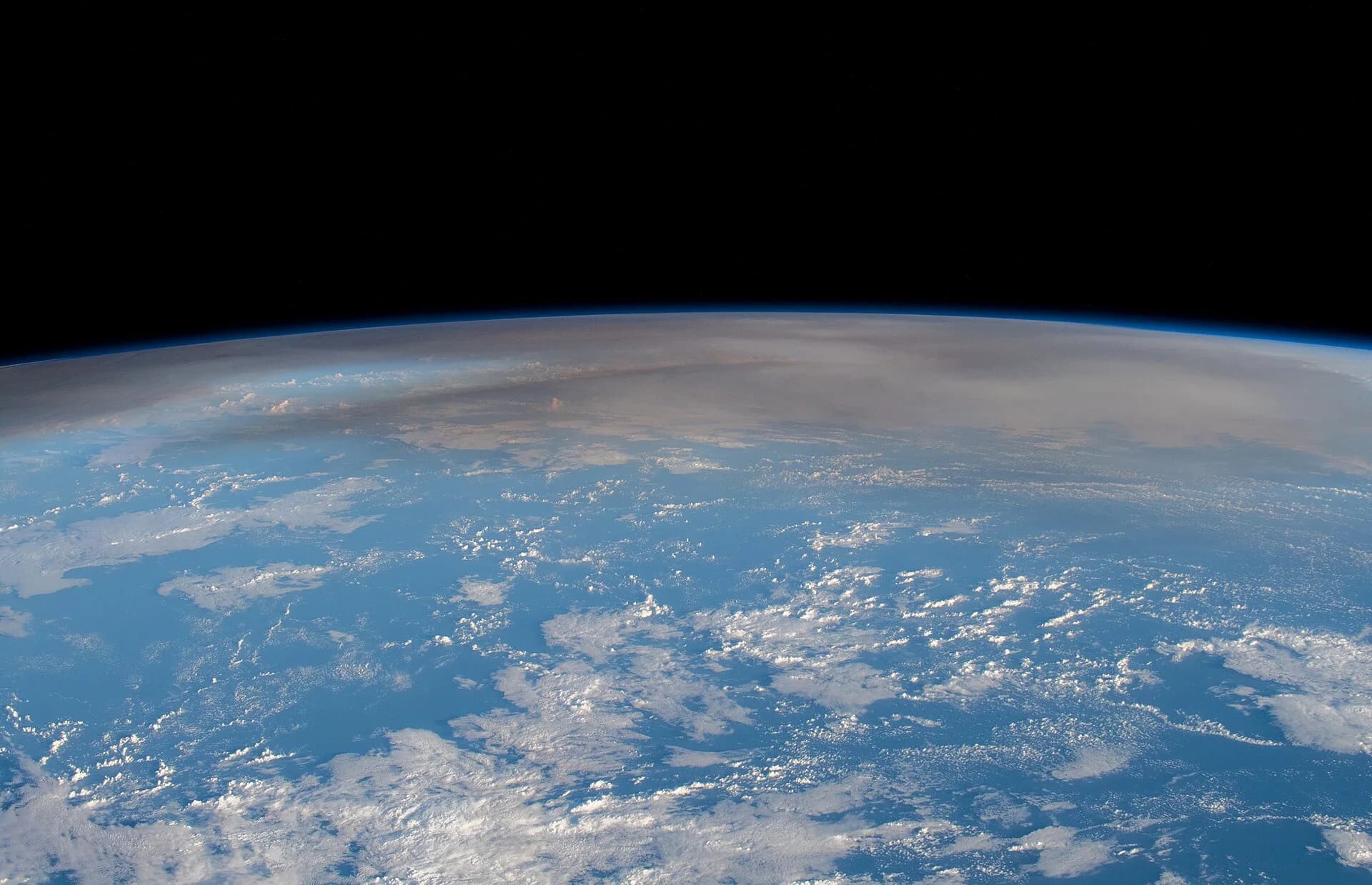 Photograph from the International Space Station on 16 January 2022, north-west of New Zealand, looking across the curved edge of the Earth: a broad, brown-grey veil of volcanic ash and gas from the Hunga Tonga eruption spreads high above white clouds over the blue Pacific Ocean.