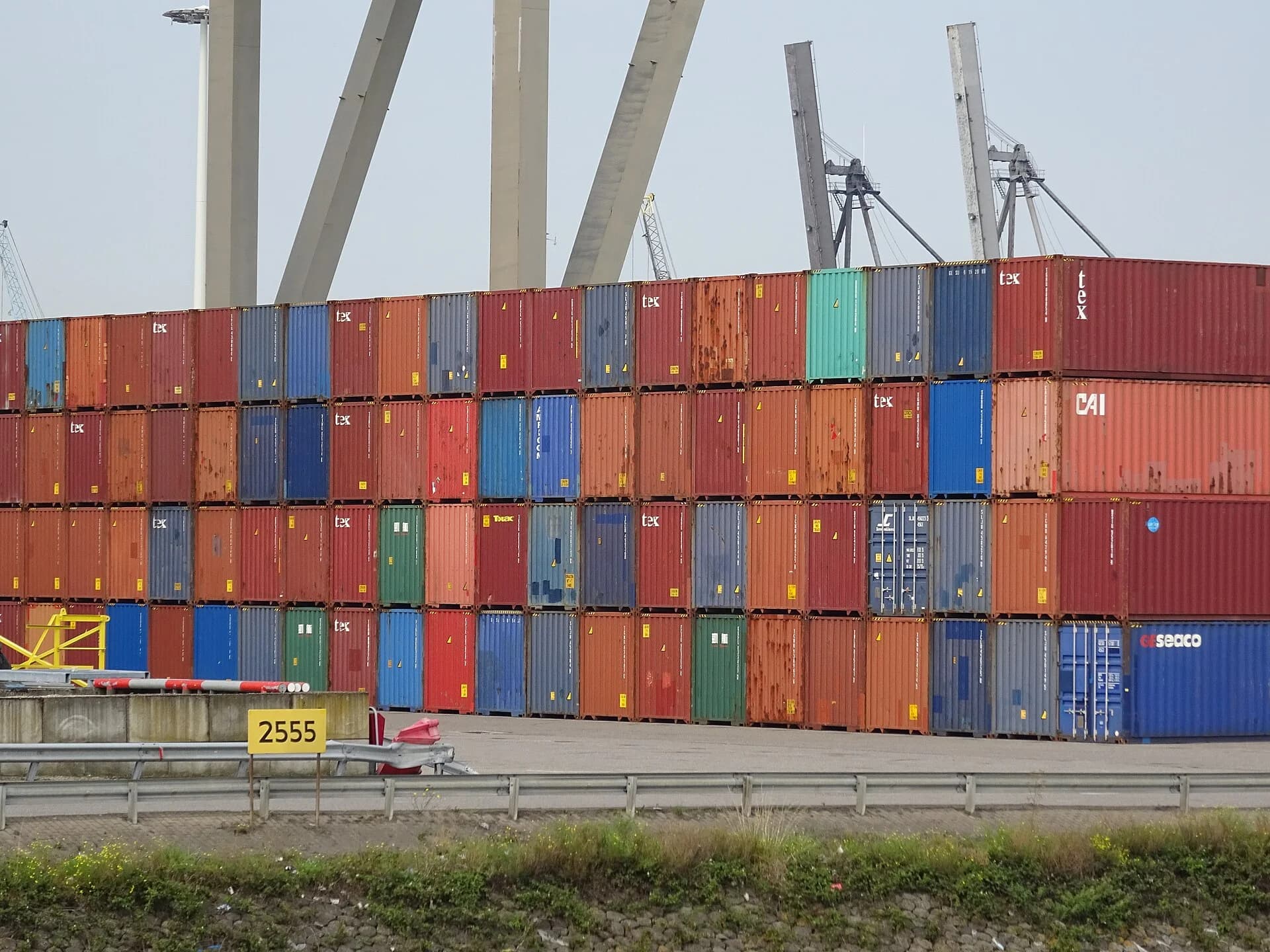 A long wall of multicoloured shipping containers stacked five high at the Port of Rotterdam, with container cranes behind.