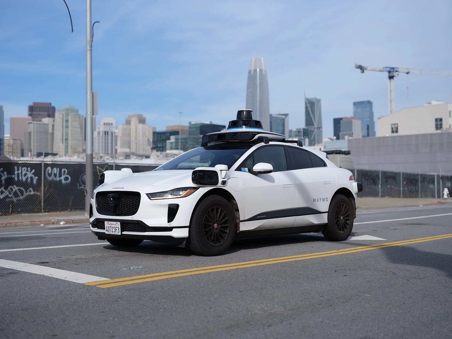 A white Waymo Jaguar I-Pace on a San Francisco street with nobody in the driver's seat, a lidar dome on the roof and sensor pods on the corners.